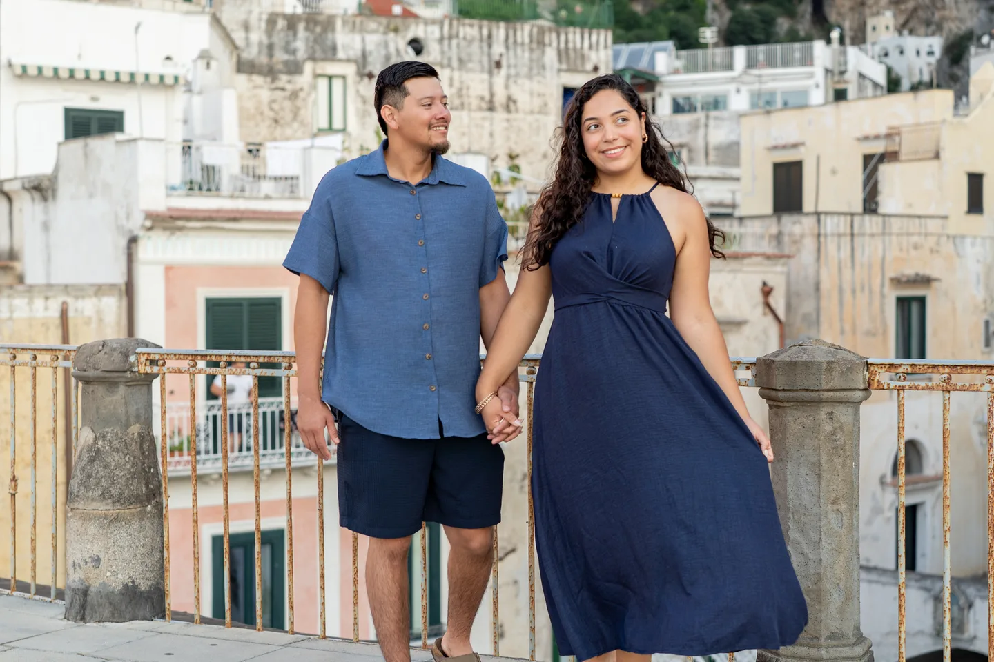 The couple smiling and holding hands by the village railing
