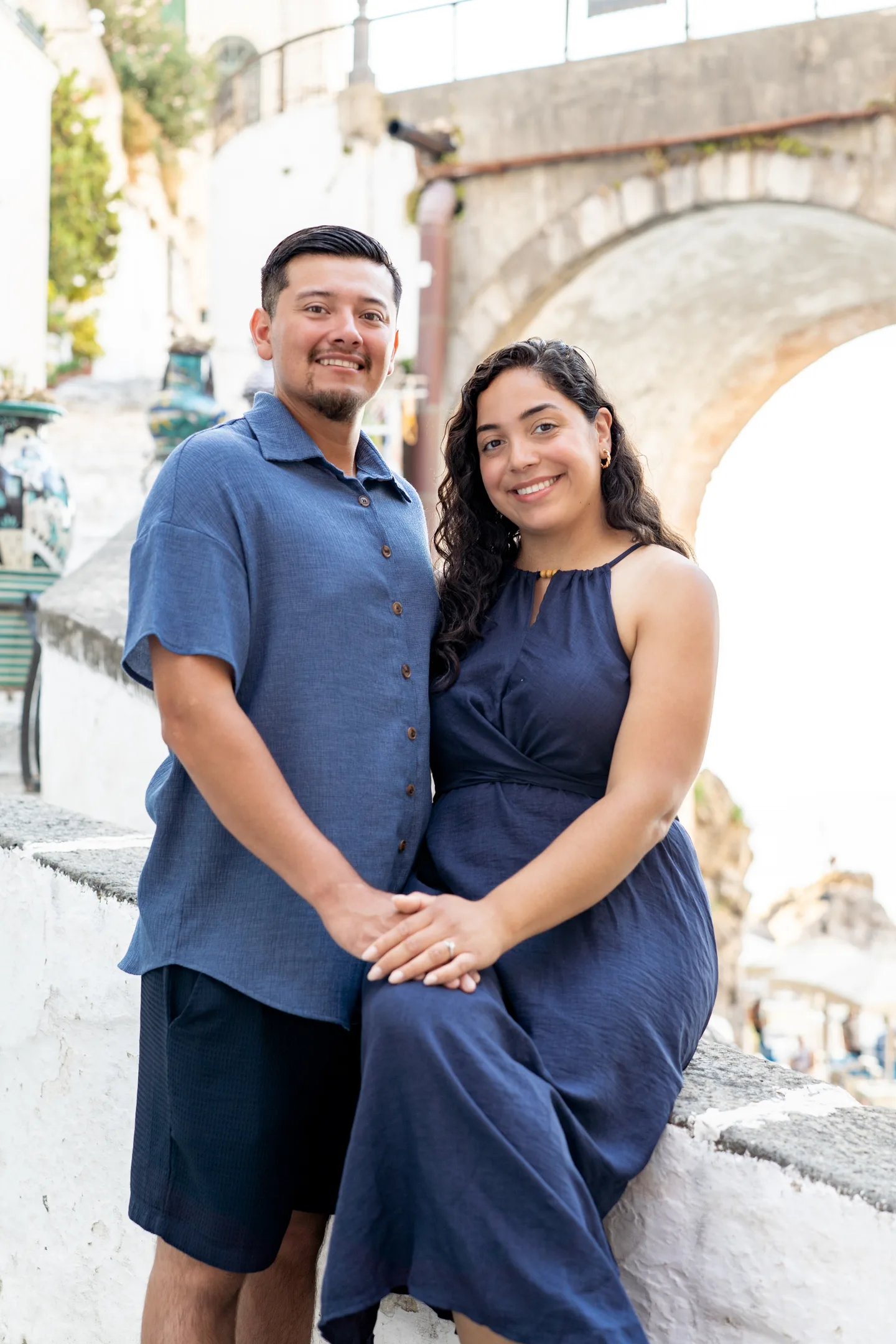The couple holding hands beside a ceramic vase and stone arch